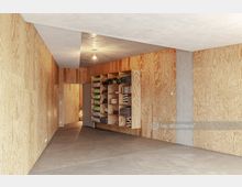 Minimalistischer Raum mit Wänden aus Sperrholz und einem Betonelement auf der rechten Seite. Links befindet sich eine Regalwand mit zusammengerollten Matten, Handtüchern und anderen Gegenständen, während der Boden aus glattem Beton besteht.