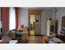 Ein Büro im Retro-Stil mit hellgemusterten Tapeten, großen Fenstern mit rosafarbenen Vorhängen und einem Kachelofen an der rechten Wand. Es gibt mehrere Schreibtische mit Akten, Schreibutensilien und einem alten Telefon, sowie eine geöffnete Tür, die in einen weiteren Raum führt.