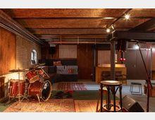 Ein Proberaum mit Backsteinwänden und Holzelementen, ausgestattet mit einem Schlagzeug, Teppichen, einer erhöhten Sitzfläche mit Kissen und einem Tresen im Hintergrund. Die Beleuchtung ist warm, und mehrere Mikrofone und Kabel sind verteilt.