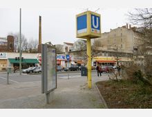 Straßenszene in einer Stadt mit einem gelben U-Bahn-Schild für die Linie U9, einer Straßenkarte in einem grauen Schaukasten und umliegenden Gebäuden, darunter Geschäfte mit bunten Fassaden. Im Hintergrund sind mehrere parkende und fahrende Autos sowie ein Backsteinschornstein sichtbar.