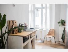 Helle Küche mit einfachem Design, einem Holzarbeitsbereich mit Schneidebrettern und einer Spüle. Im Hintergrund ein rundes Tischchen, ein geflochtener Stuhl und eine große Fensterfront mit weißen Vorhängen; Zimmerpflanzen und Holzboden ergänzen den Raum.