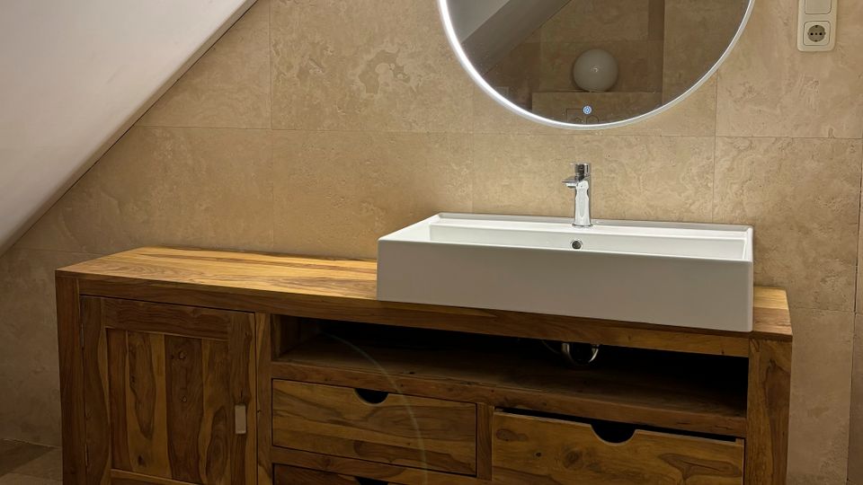 Modernes Badezimmer mit schräger Dachwand, beigefliesten Wänden und Boden sowie einem langen Holzwaschtisch mit Schubladen und rechteckigem Aufsatzwaschbecken. Darüber hängt ein runder, beleuchteter Spiegel; rechts ist eine Steckdose an der Wand zu sehen.