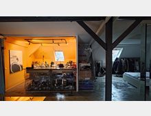 Ein Dachgeschossraum mit offener Küche auf der linken Seite, gekennzeichnet durch orange Wände, ein kleines Dachfenster und eine Arbeitsplatte mit Haushaltsgegenständen. Rechts davon ein Schlafbereich mit einem Bett, Kleiderständern und einem weiteren Dachfenster; dunkle Holzbalken trennen die Bereiche.