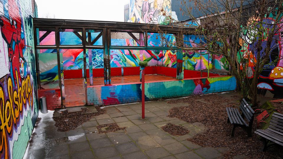 Innenhof mit gepflastertem Boden und Sitzbänken, umgeben von Wänden mit großflächigen, bunten Graffiti-Motiven. In der Mitte steht eine offene, überdachte Plattform mit Holz- und Glasrahmen sowie niedriger, farbig gestrichener Brüstung; daneben ein kahler Baum.