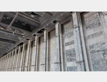 Innenansicht eines Gebäudes im Rohbauzustand mit einer Wand aus Beton- und Steinplatten, verstärkt durch freiliegende vertikale Betonsäulen und Metallgerüste. Die Decke zeigt sichtbare Metallstreben und Öffnungen.