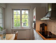 Helle, schmale Küche mit weißer Einbauzeile und Holzarbeitsplatte, Cerankochfeld sowie Edelstahl-Dunstabzugshaube an einer gefliesten Wand. Ein großes Sprossenfenster mit Blick ins Grüne und ein kleiner Esstisch am Fenster prägen den Raum.