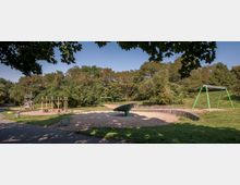 Spielplatz mit mehreren Spielgeräten, darunter eine Schaukel, ein Klettergerüst aus Holz und ein drehbares Karussell auf einem Sandbereich, umgeben von Rasenflächen und Bäumen. Im Hintergrund dichter Baumbestand und ein klarer Himmel.