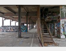 Ein leerstehendes, verlassenes Gebäude mit Betonwänden, Stahlträgern und einer Holzwendeltreppe, dessen Wände und Oberflächen mit Graffiti bedeckt sind. Der Raum wirkt ungenutzt und verwittert, mit Schutt auf dem Boden und leerem, lichtdurchflutetem Hintergrund.