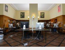 Ein heller Arbeitsraum mit hohen Decken, Holzwandverkleidung und Regalen voller Bücher. Mehrere Schreibtische mit Computern (iMacs) und Schreibtischlampen sind auf dem karierten Fliesenboden angeordnet, während Personen an einigen Stationen arbeiten.