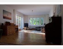 Helles Wohnzimmer mit Parkettboden, großen Fenstern und Blick auf einen Garten. Der Raum ist möbliert mit einem grauen Sofa, einem kleinen Couchtisch, einem Fernseher, einem Schreibsekretär und einer Kommode mit vielen Schubladen.