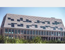 Modernes Gebäude mit einer Fassade aus horizontalen Holzpaneelen und unregelmäßig angeordneten Fenstern im oberen Teil. Die untere Etage hat große, symmetrische Glasfenster, und im Vordergrund sind Gräser zu sehen.