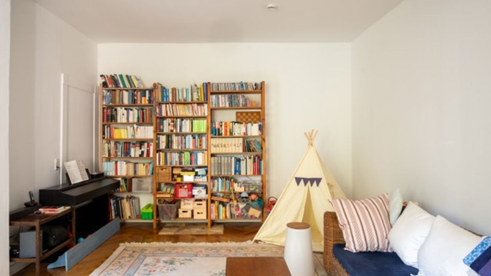 Wohnzimmer mit Holzfußboden, großem Bücherregal an der Rückwand und einem Klavier an der linken Wand. Rechts stehen ein geflochtener Sofa mit Kissen, ein kleiner Couchtisch und ein Tipi-Zelt auf einem gemusterten Teppich.