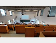 Ein Hörsaal mit ansteigenden Sitzreihen aus Holz, einer breiten Tafel und einem Dozentenpult vorne. Der Raum ist hell, mit großen Fenstern an den Seitenwänden und einer weißen Projektionsfläche an der Frontwand.