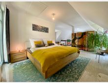 Modernes Schlafzimmer mit einem großen Holzbett, gelber Bettdecke und dekorativen Kissen. Der Raum enthält einen Nachttisch mit Lampe, einen Raumteiler mit Waldmotiv und eine große Zimmerpflanze; helle Wände und ein Fenster links lassen viel Licht hinein.