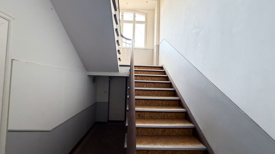 Innenansicht eines Treppenhauses mit gerader Treppe, braun gemusterten Stufen und dunkelbraunem Handlauf, die zu einem Podest führt. Helle Wände mit grauem Sockelbereich und ein großes Fenster am Treppenabsatz lassen Tageslicht in den Raum.