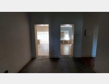 Blick in einen dunklen Flur mit zwei offenen Türrahmen, die in zwei helle, leerstehende Räume führen. Beide Räume haben große Fenster und sichtbare Heizkörper unter den Fenstern, der Boden besteht aus Parkett.