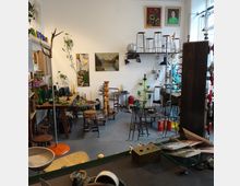 Ein kleiner, eklektischer Laden mit Vintage-Möbeln, Glaswaren, Kerzenhaltern und dekorativen Gegenständen. An den Wänden hängen Gemälde, auf Regalen und Tischen sind verschiedene Retro-Objekte ausgestellt.