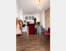 Innenraum eines Studios oder Ateliers mit dunklem Holzboden, offener Treppe zu einer Empore und weißem Geländer; darüber hängt ein kleiner Kronleuchter. Links steht ein Kleiderständer mit Kostümen, mittig trennt ein roter Vorhang den Bereich ab, rechts sind eine Betonwand und eine rote Sitzbank zu sehen.