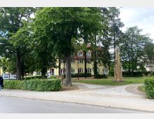 Grüne Parkanlage mit gepflasterten Wegen, umgeben von Bäumen und Büschen, im Hintergrund ein mehrstöckiges gelbes Gebäude mit roten Dachziegeln und großen Fenstern. Rechts steht eine moderne Steinskulptur mit einer kleinen Uhr an der Spitze.