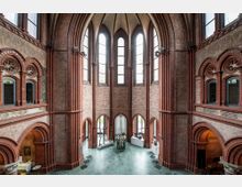 Innenraum eines neugotischen Gebäudes mit hohen Spitzbogenfenstern, roten Backsteinwänden und verzierten Bögen. Der Raum zeigt eine Ausstellung mit Skulpturen auf weißen Podesten und verfügt über einen grünen Marmorboden.