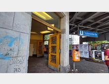 U-Bahn-Station „Schlesisches Tor“ in Berlin mit einem offenen Eingangsbereich, gelben Holztüren mit Glaseinsatz, grauem Kachelboden und einer ausgeschilderten blauen U-Bahn-Stationstafel. Im Hintergrund sind Plakate an einer Wand sowie ein orangefarbener Mülleimer zu sehen.