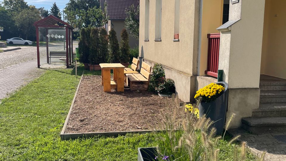 Außenansicht eines hell verputzten Gebäudes an einer ruhigen Straße, mit kleinem Sitzbereich aus Holzbänken und Tisch auf einem Beet neben dem Eingang. Im Vordergrund stehen Pflanzkübel mit Ziergräsern und lila Blumen; links ist eine verglaste Bushaltestelle am Straßenrand zu sehen.