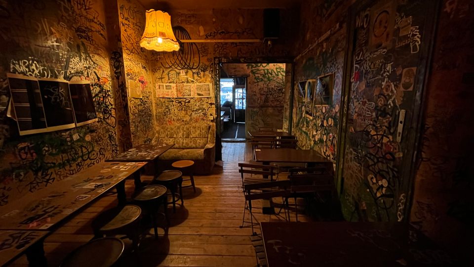 Schmaler, schwach beleuchteter Innenraum einer Bar oder Kneipe mit Holzboden, einfachen Tischen und Hockern sowie einer hängenden Lampe. Die Wände und Türen sind dicht mit Graffiti, Stickern und Kritzeleien bedeckt, im Hintergrund führt ein Durchgang in einen weiteren Raum.