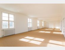 Ein leerer, heller Raum mit beigen Teppichboden, weißen Wänden und großen Fenstern auf der linken Seite, die mit Heizkörpern darunter ausgestattet sind. Der Raum hat eine rechteckige Form und endet an einer Wand mit zwei offenen Nischen.