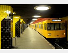 U-Bahn-Station mit gelben und schwarzen Kacheln an den Säulen, hellem Boden und einer gewölbten Decke mit runden Leuchten. Ein gelber Zug der Linie U7 mit der Anzeige "Rathaus Spandau" steht am Bahnsteig.