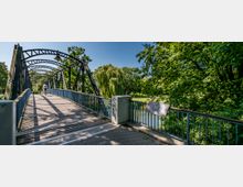 Eine historische Stahlbogenbrücke mit Holzboden führt über einen kleinen Fluss und ist von üppigem, grünem Baumbestand umgeben. Im Vordergrund steht eine Infotafel am Brückengeländer, und im Hintergrund sind ein Radfahrer sowie eine parkähnliche Landschaft mit einer Trauerweide zu sehen.