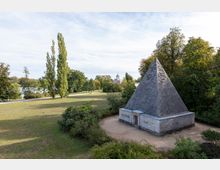 Steinerne Pyramide in einer parkähnlichen Landschaft, umgeben von Bäumen und Rasenflächen; im Hintergrund ist ein Fluss sowie ein historischer Kuppelbau erkennbar.