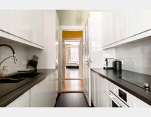 Schmale, moderne Küche mit weißen, glänzenden Schränken, schwarzer Arbeitsplatte und Fliesenrückwand in Weiß. Im Hintergrund offene Tür mit Blick auf ein Schlafzimmer mit Holzboden und großem Fenster.