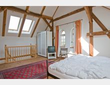 Dachgeschoss-Schlafzimmer mit sichtbaren Holzstützbalken, zwei Dachfenstern, weißen Wänden und einem roten Teppich. Der Raum enthält ein Bett, einen Kleiderschrank, einen kleinen Fernseher auf einem Beistelltisch, einen Stuhl und Holzboden, mit Zugang zu einer Treppe.