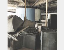 Innenraum einer unvollendeten oder renovierungsbedürftigen Industriewerkhalle mit freiliegenden Deckenstrukturen und Metallgerümpel im Vordergrund, einschließlich Rohre, Bleche und anderer Baustoffe. Im Hintergrund befindet sich eine große, zylindrische, blau-graue Struktur.