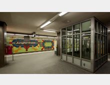 U-Bahnhof Jungfernheide mit einer bunten Mosaikwand und dem Stationsnamen in der Mitte, daneben ein gläserner Aufzugsschacht mit Metallrahmen. Die Bahnhofshalle ist modern beleuchtet und zeigt klare architektonische Linien.