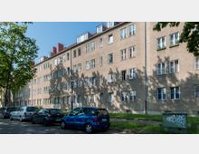 Mehrstöckiges Wohngebäude mit schlichtem Putzfassadenstil und zahlreichen Fenstern in regelmäßiger Anordnung, umgeben von geparkten Autos und vereinzelten Bäumen, die Schatten auf die Fassade werfen. Im Vordergrund ein Gehweg und ein Stromkasten mit Graffiti.