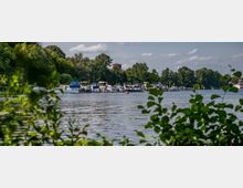 Flusslandschaft mit einer Reihe von Booten und Yachten, die an einem Steg angelegt sind, umgeben von dicht bewachsenen Ufern mit Bäumen; im Hintergrund ist ein kleiner Ziegelturm sichtbar.