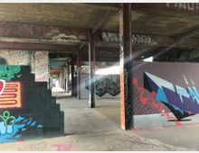 Industriehalle mit Betonwänden und -säulen, bedeckt mit bunten Graffiti und Wandgemälden. Der Boden ist aus Beton, und Lichtstrahlen dringen durch offene Stellen in der Gebäudestruktur.