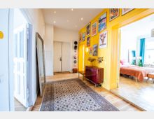 Flur einer Wohnung mit gelben Wänden, einem großen Spiegel, einer weißen Doppeltür und einem verzierten Teppich auf Holzboden. An der Wand hängen bunte Kunstwerke, daneben befindet sich ein rotes Sideboard; rechts führt eine offene Tür in ein Schlafzimmer mit einem Bett und türkisfarbenen Vorhängen.