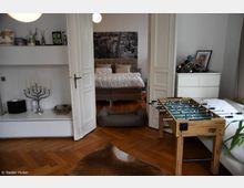 Ein Wohnzimmer mit hölzernem Fischgrätparkett, das über eine Doppeltür in ein Schlafzimmer führt. Im Vordergrund stehen ein Kickertisch, ein Hundebett und ein Teppich; links sind Regale mit Dekorationsgegenständen, rechts eine Kommode mit Weinregal und Fotos.