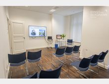 Kleiner Konferenzraum mit mehreren blauen Stühlen, die in Reihen angeordnet sind, einem großen Bildschirm an der Wand mit einer Begrüßungsanzeige, einem kleinen Tisch mit Blumen und einer Lampe sowie einem Fenster mit vertikalen Jalousien. Der Raum hat einen Holzboden und helle Wände.