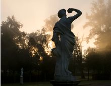 Eine klassische Marmorskulptur im Vordergrund steht in einem Park, beleuchtet von diffusem Licht bei Sonnenaufgang oder -untergang. Im Hintergrund sind Bäume und eine weitere Statue in der Ferne zu sehen, umgeben von Nebel und warmem Licht.