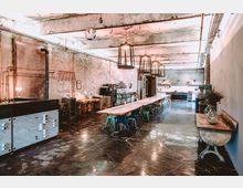 Industriell gestalteter Raum mit offener Küche, langer Holztischgruppe und bunten Stühlen. Sichtbare Backsteinwände, freiliegende Rohrleitungen und große Lampen bestimmen das Ambiente.