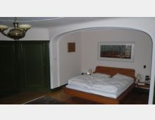 Ein Schlafzimmer mit einem Holz-Doppelbett, weißer Bettwäsche und zwei Nachttischen mit Lampen. Der Raum hat grüne Einbauschränke, einen gewölbten Durchgang und ein Landschaftsbild an der Wand.