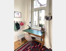 Helle Ecke eines Kinderzimmers mit einem Holzschreibtisch und grüner Arbeitsfläche, davor ein grauer Stuhl. Im Hintergrund ein großes Fenster mit weißen Vorhängen, an der Wand bunte Taschen und ein Bild, auf dem Boden ein farbig gemusterter Teppich.