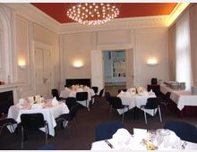 Eleganter Speiseraum mit weiß gedeckten Tischen, dunkel gepolsterten Stühlen und kunstvollen Stuckwänden. An der Decke hängt ein moderner Kronleuchter, und ein Buffetbereich ist an der rechten Wand eingerichtet.