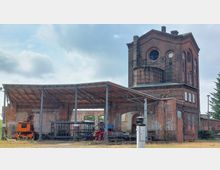 Ehemaliges Backsteingebäude mit industrieller Architektur, bestehend aus einem hohen Turm mit Rundfenstern und einem angebauten offenen Unterstand mit Metallträgern und Wellblechdach. Im Vordergrund stehen ein orangefarbener Kran und verschiedene alte Industriemaschinen.