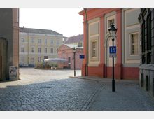 Gepflasterter Platz in einer historischen Stadt mit pastellfarbenen Gebäuden, darunter ein gelbes Haus und ein rosa Gebäude mit klassizistischen Fassaden. Im Vordergrund stehen eine Straßenlaterne und ein Schild für barrierefreies Parken.
