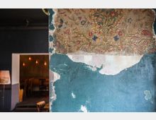 Eine Nahaufnahme einer Wand mit abgeblätterter Farbe; der obere Bereich zeigt eine verblasste, florale Tapete mit goldenen, roten und blauen Mustern, während der untere Bereich mit blauer Farbe gestrichen und teilweise beschädigt ist. Im Hintergrund ist durch eine Türöffnung ein Raum mit Holzstühlen und warmem Licht sichtbar.