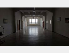 Ein langer, leerer Flur mit glänzendem Boden und weißen Wänden, beleuchtet von einer großen Fensterfront am Ende. Entlang der Seitenwände befinden sich einige Türen sowie Wanddekorationen.